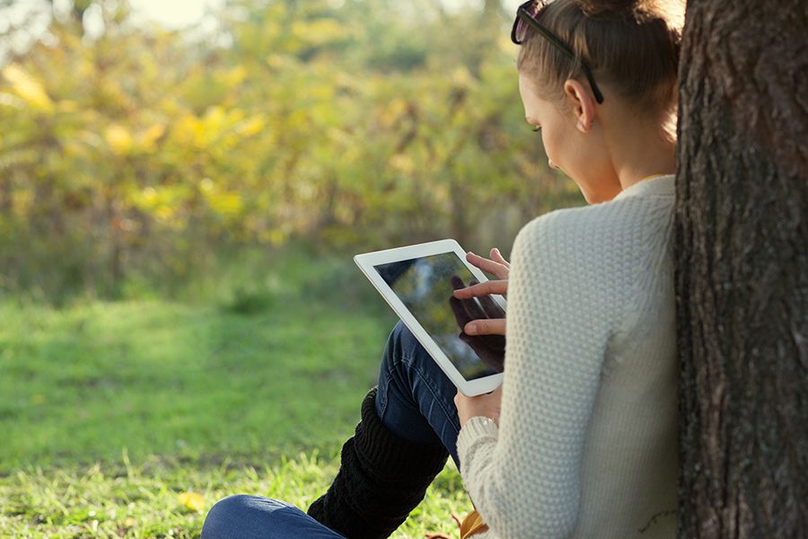 Client Center - Colorful Close Up of Young Woman Sitting and Leaning Against a Tree While Accessing Account Information on Her Tablet