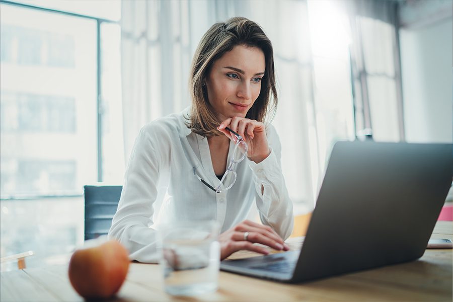 UPMC Member Resources - Professional Woman Smiling and Thinking While Scrolling on Her Laptop at Her Desk with a Large Window Behind Her Pouring in Light